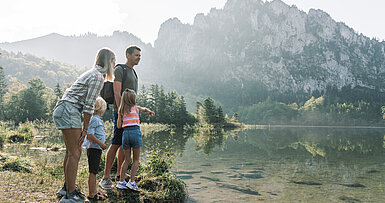 Eine Familie steht am Seeufer des Laudachsee in Grünberg im Salzkammergut und schaut aufs Wasser, die Berge spiegeln sich im See, (c) Oberösterreich Tourismus/Traunsee-Almtal/Robert Maybach